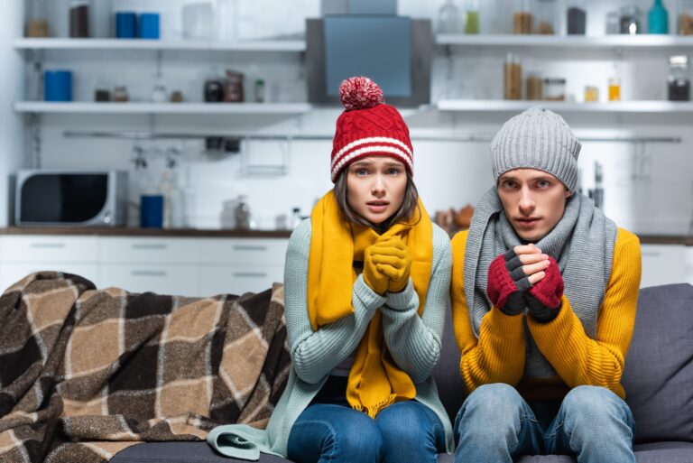 freezing couple in warm hats and gloves looking at camera while sitting on sofa in cold kitchen