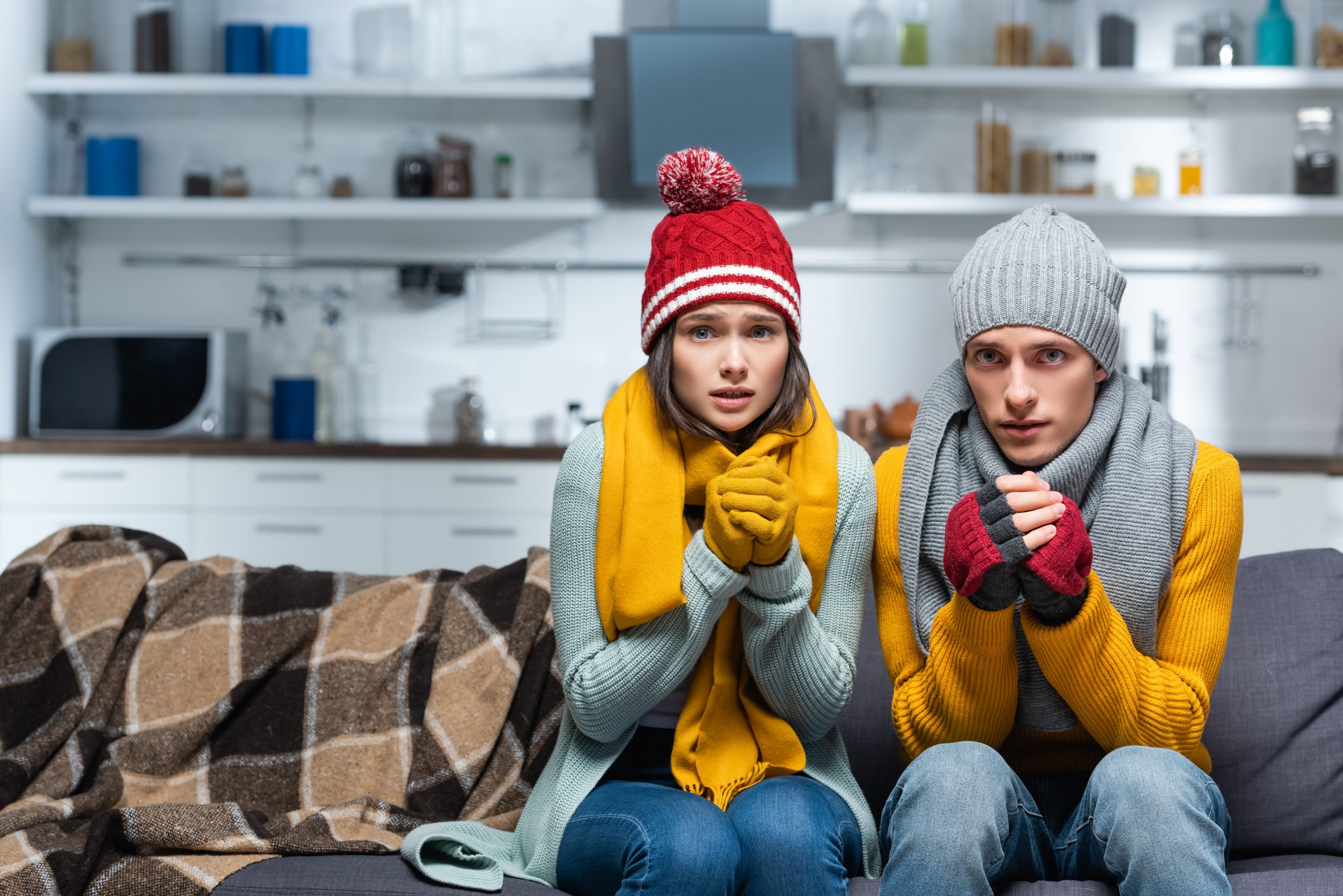 freezing couple in warm hats and gloves looking at camera while sitting on sofa in cold kitchen