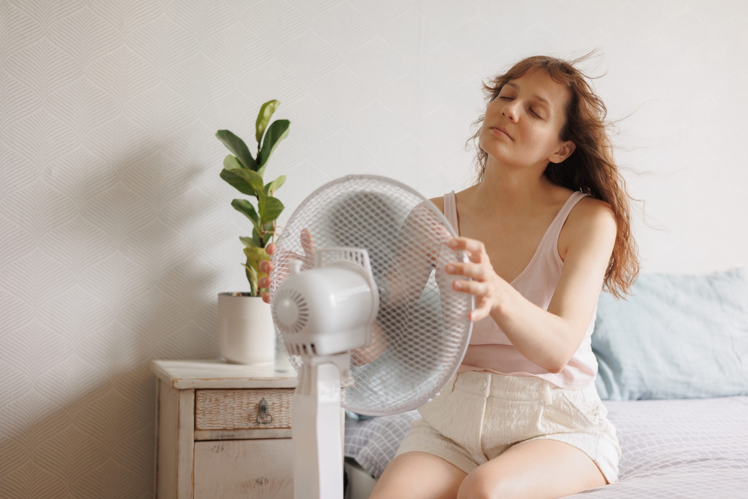 woman cools down at home with help of an air conditioner fan, sits on bed in front of an electric